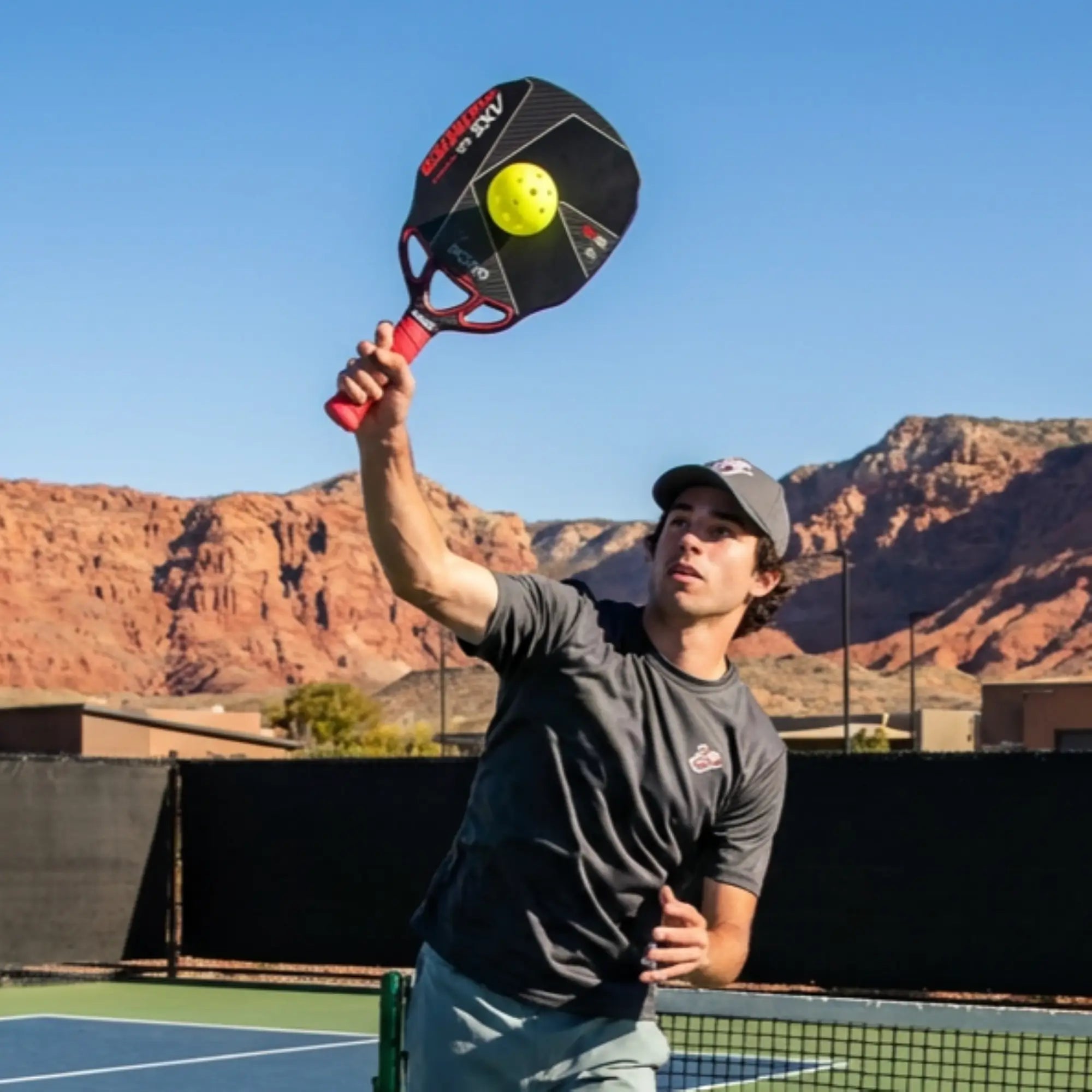 Person playing pickleball on a court with mountains in the background with KOBO Thunder AXE Infinity pickleball paddleon hand doing a overhead mash.